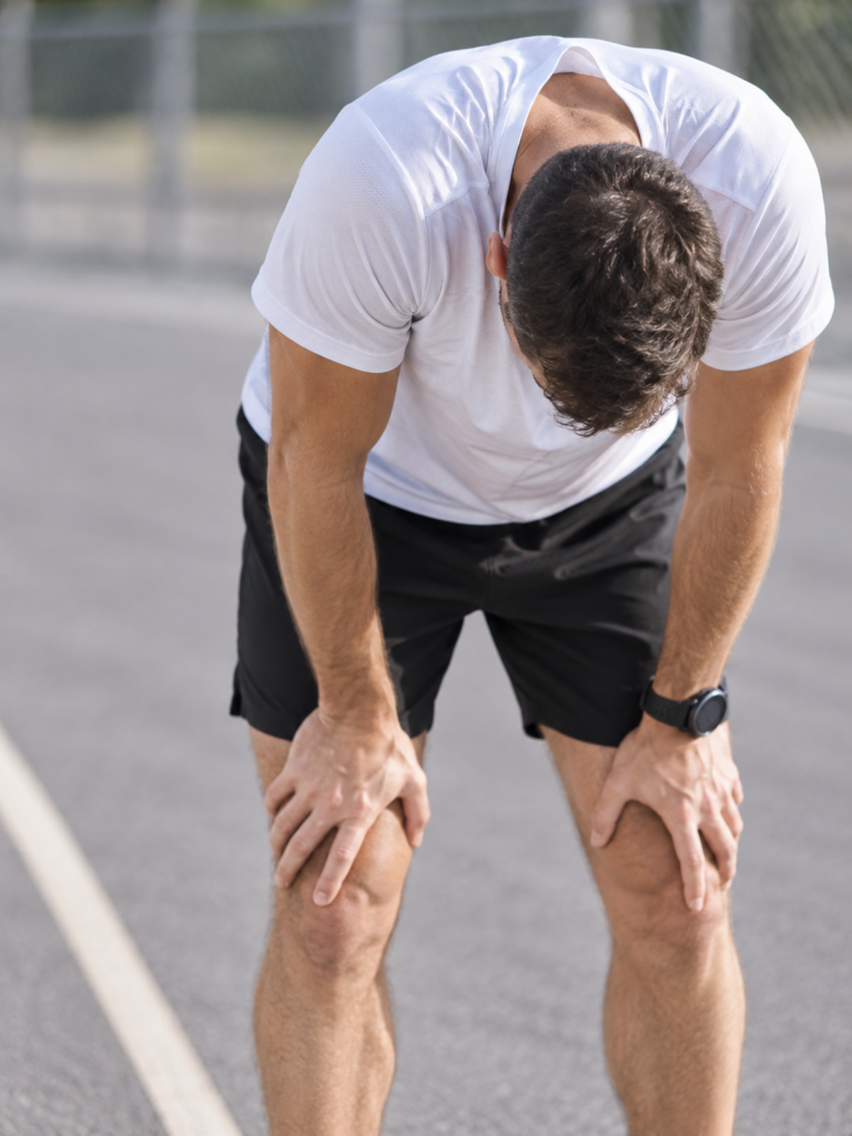 Man in a white athletic shirt resting with his hands on his knees after a run.